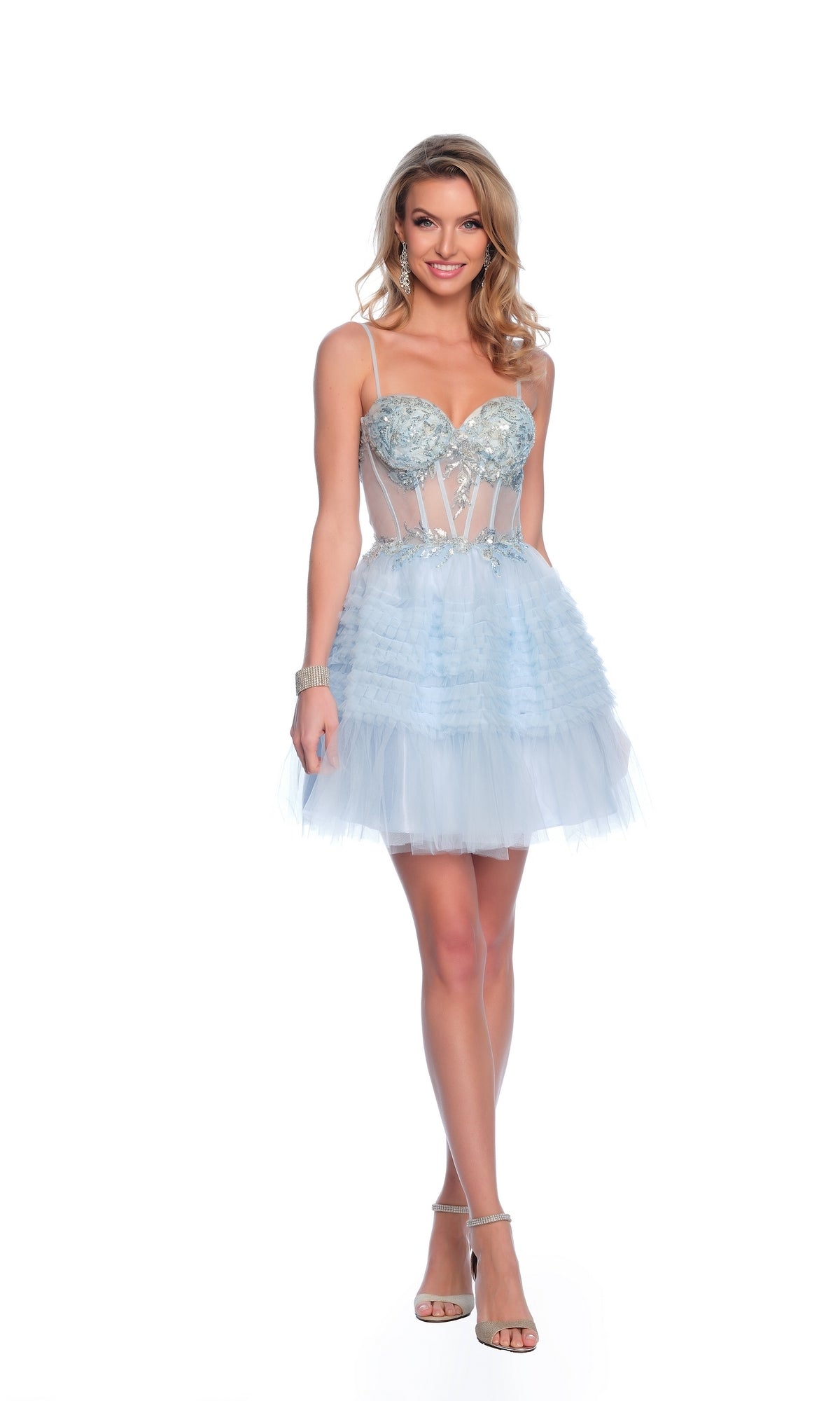 A woman wears the Dave & Johnny Short Blue Ruffled Hoco Dress 11888, a strapless, light blue mini dress with layered tulle, sheer panels, and a corset bodice, standing and smiling against a white background.
